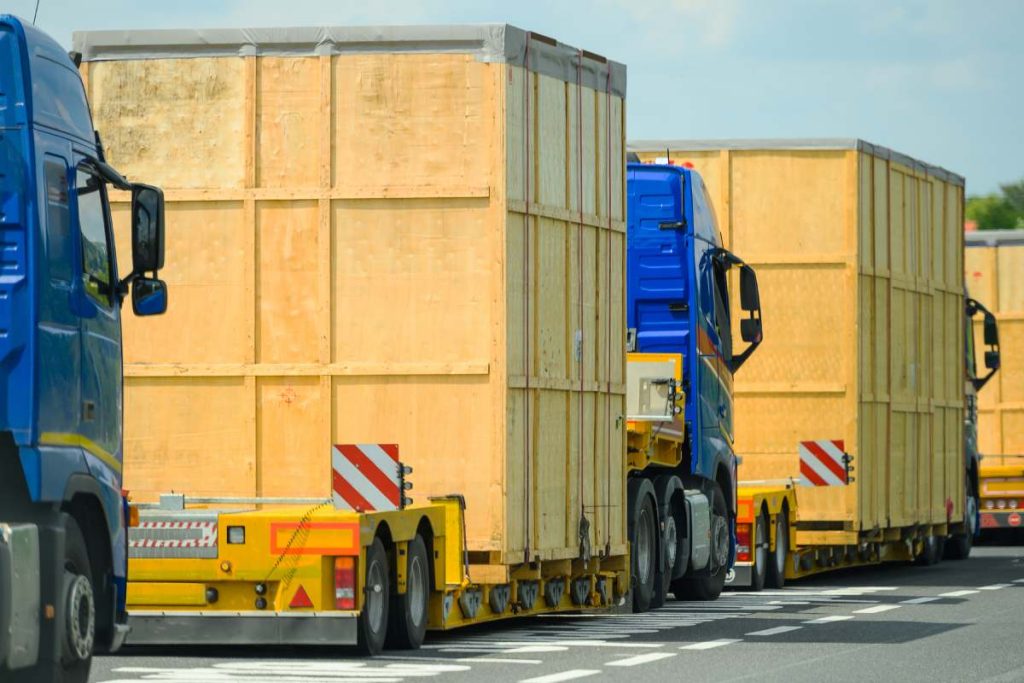 Heavy Oversize Load Convoy on the Route to Destination. Few Semi Trucks Pulling Large Wooden Oversized Containers. Transportation Industry.